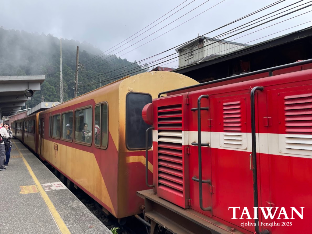 A close-up view of a bright red Alishan Forest Railway locomotive and its cream-and-red passenger carriages stopped at the Fenqihu Station platform under a cloudy, misty sky.