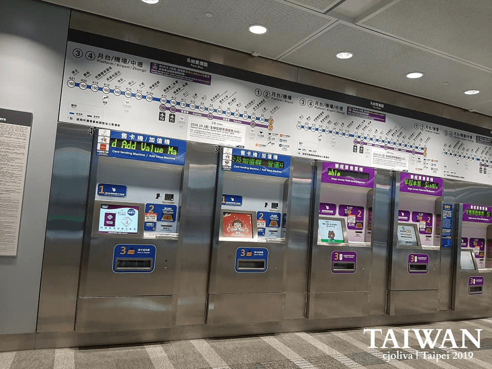 A row of modern stainless steel ticket vending and card add-value machines at a Taoyuan Airport MRT station in Taipei, Taiwan. Above the machines is a large, detailed system map showing routes to the airport and Zhongli, with clear fare information.
