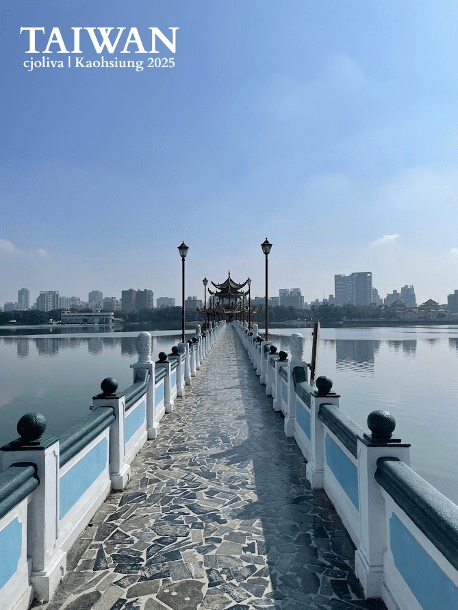 A long, symmetrical stone bridge with blue and white railings leading to a traditional Chinese pavilion on a calm lake in Kaohsiung, Taiwan.
