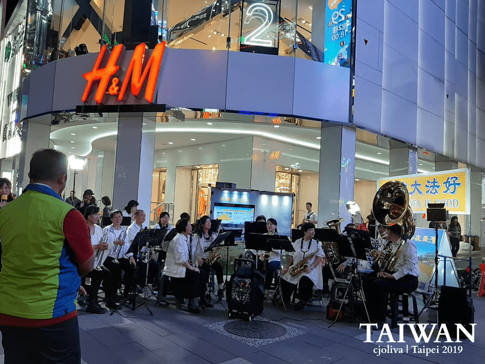 Musicians performing in Ximending, Taipei, Taiwan, with brass and woodwind instruments in front of an H&M store, accompanied by signage and a lively urban backdrop, taken in 2019
