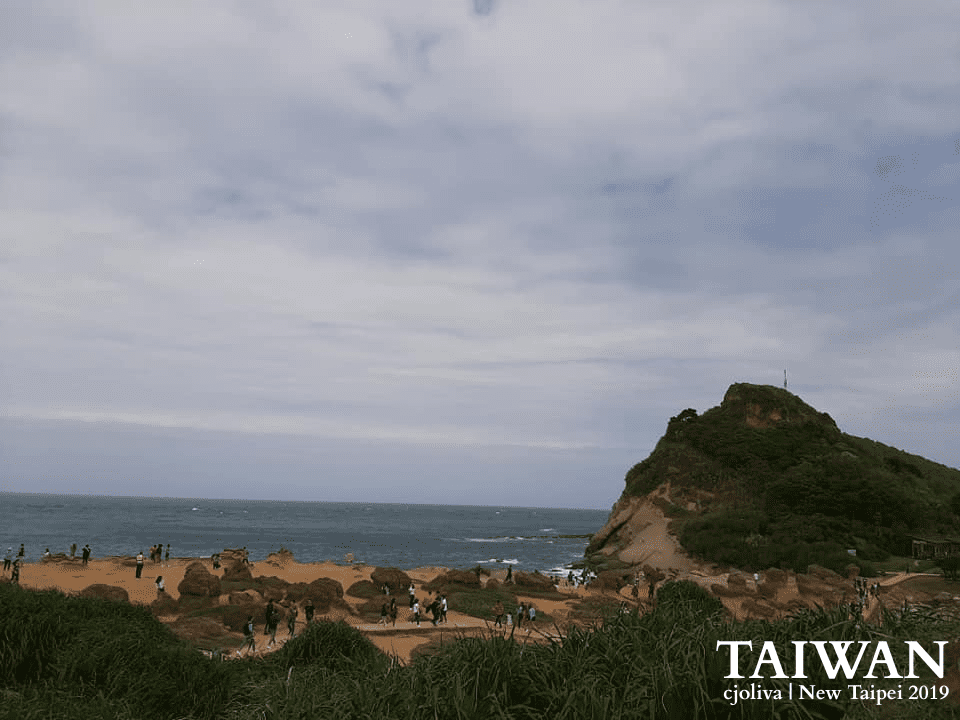Scenic coastal view near Yehliu in New Taipei, Taiwan, showing grassy terrain, rocky beach with visitors, green hill, and ocean horizon under partly cloudy skies, taken in 2019