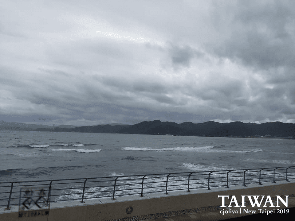 Overcast coastal scene near Yehliu Park in New Taipei, Taiwan, showing ocean waves, forested hills, and a concrete promenade with railing, taken in 2019