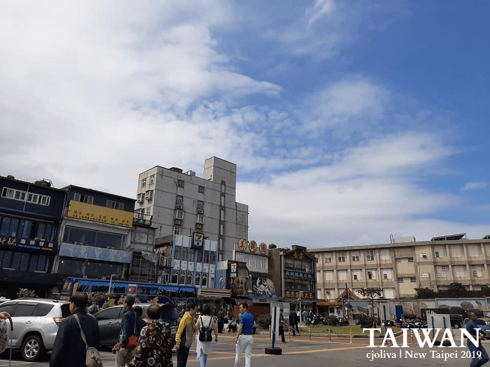 Bustling Yehliu street in New Taipei, Taiwan, with people walking near shops and a 7-Eleven under a clear sky, taken in 2019