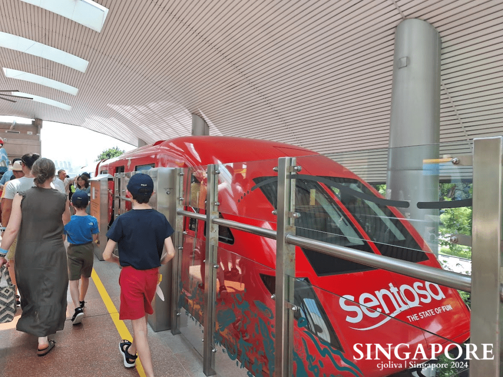 A bright red Sentosa Express monorail train stopped at a station platform with passengers boarding in Singapore.