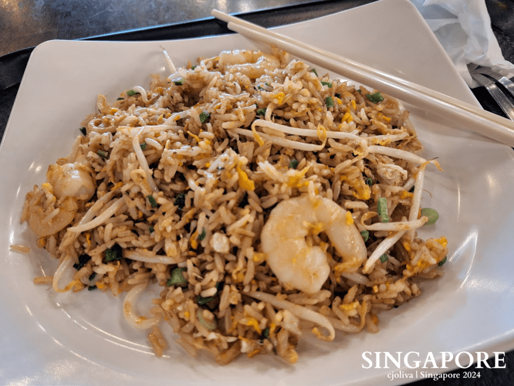 A plate of stir-fried rice with large shrimp, bean sprouts, and egg, served with white chopsticks.