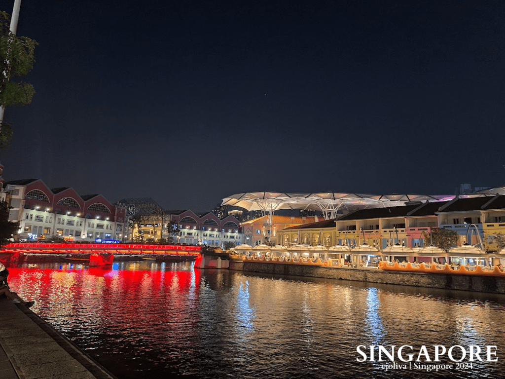 Nighttime view of Clarke Quay in Singapore with colorful illuminated buildings, arched roofs, canopy structures, and reflections of red and yellow lights on the calm river.