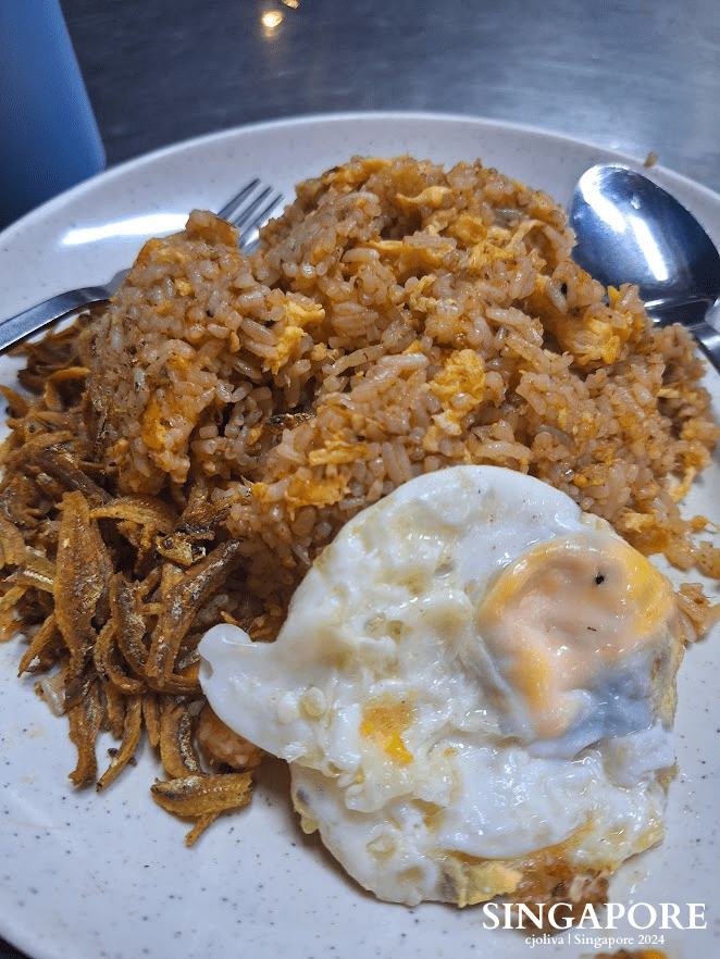 A plate of fried rice with chili seasoning, a fried egg with a runny yolk, and crispy golden anchovies beside the rice, served casually in Singapore.