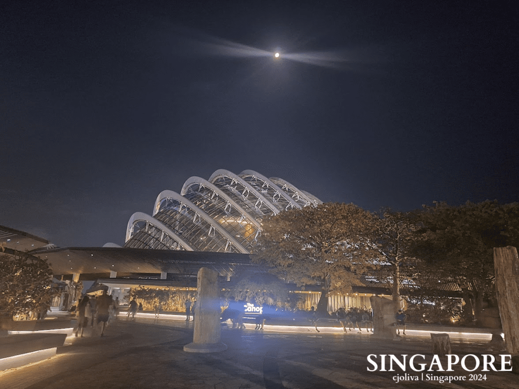 Night view of Gardens by the Bay in Singapore with illuminated glass-domed Flower Dome, moon above, trees, pathways, and people walking under warm lights.