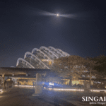 Night view of Gardens by the Bay in Singapore with illuminated glass-domed Flower Dome, moon above, trees, pathways, and people walking under warm lights.