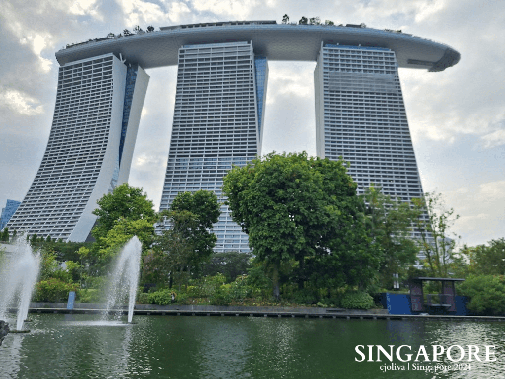 The Marina Bay Sands hotel in Singapore has three towers connected by a ship-like rooftop, a pond with fountains, and lush greenery in the foreground.