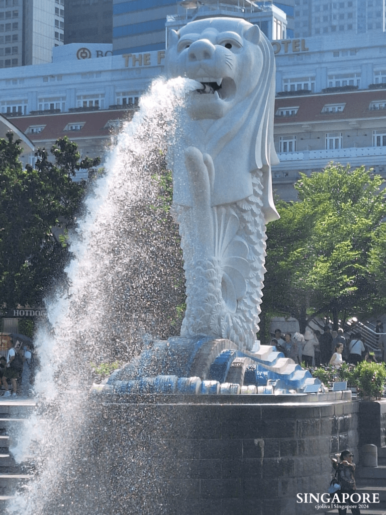 The Merlion statue in Singapore is spouting water into the bay, with The Fullerton Hotel and modern buildings in the background, surrounded by greenery and visitors.
