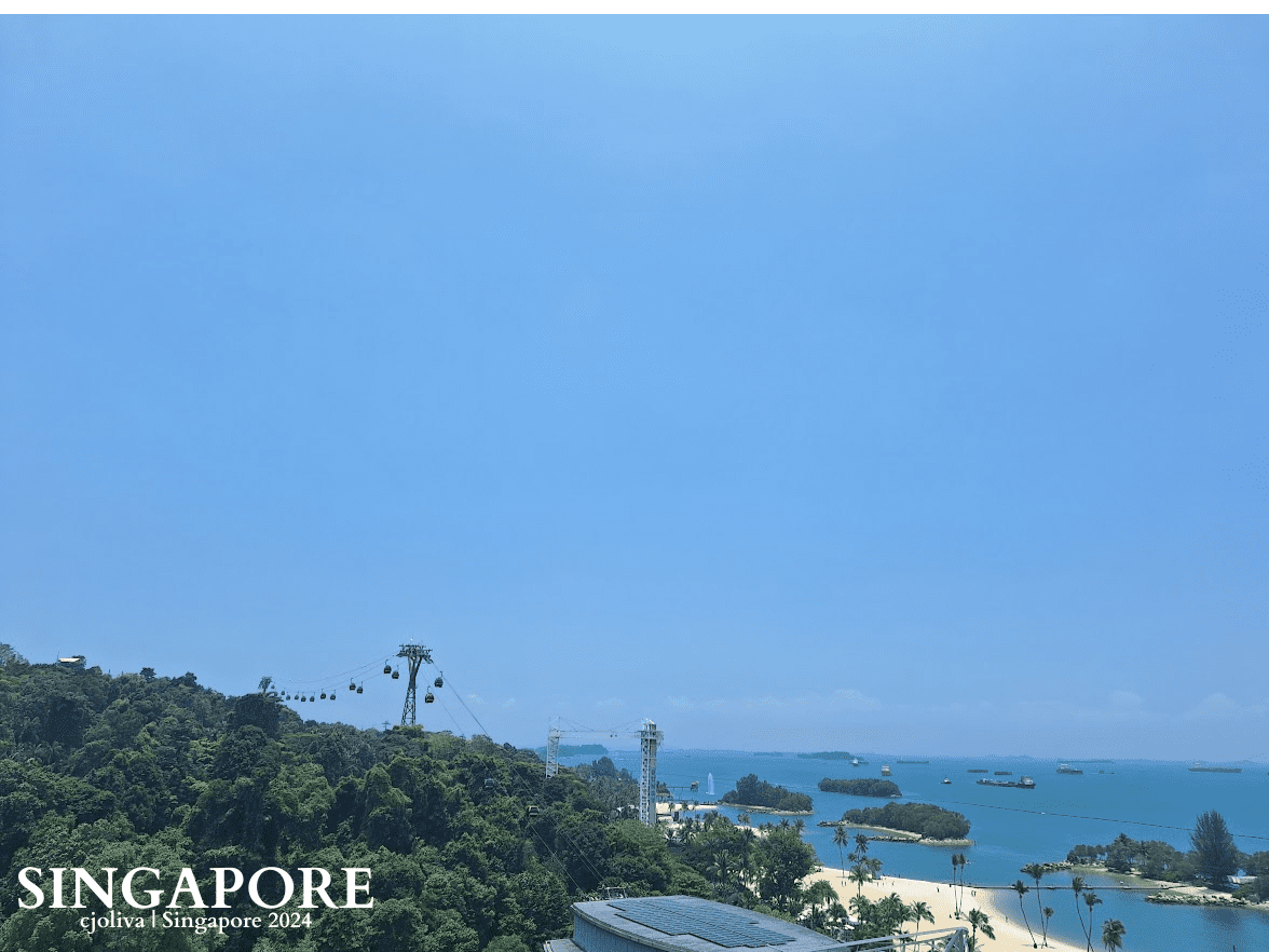 Scenic coastal view at Sentosa, Singapore, with lush green hills, sandy beaches, tropical trees, and cable cars suspended between towers over the ocean under a bright blue sky.