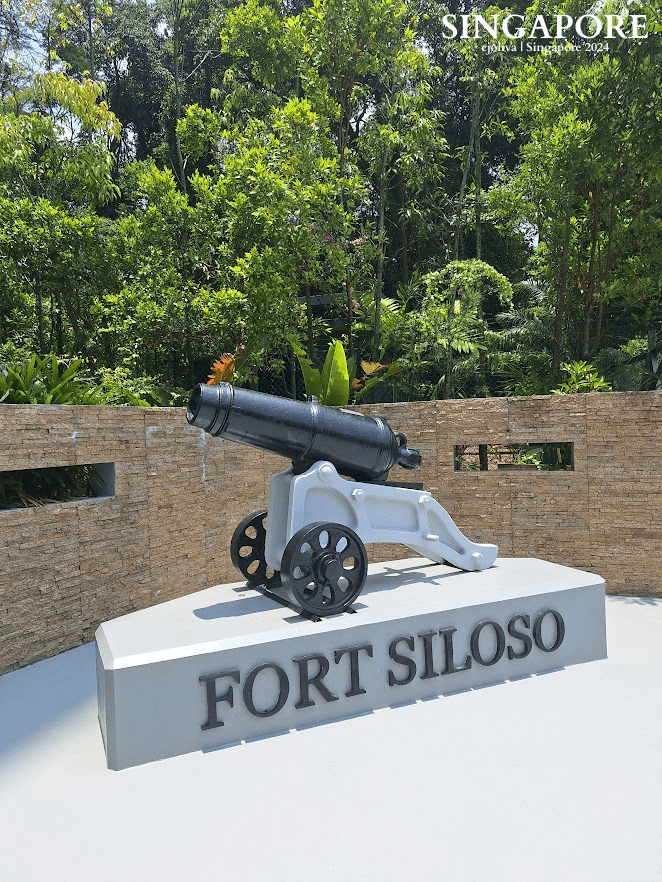 Historical cannon monument at Fort Siloso, Sentosa, Singapore, mounted on a pedestal with a “FORT SILOSO” inscription, stone wall backdrop, and lush tropical greenery.