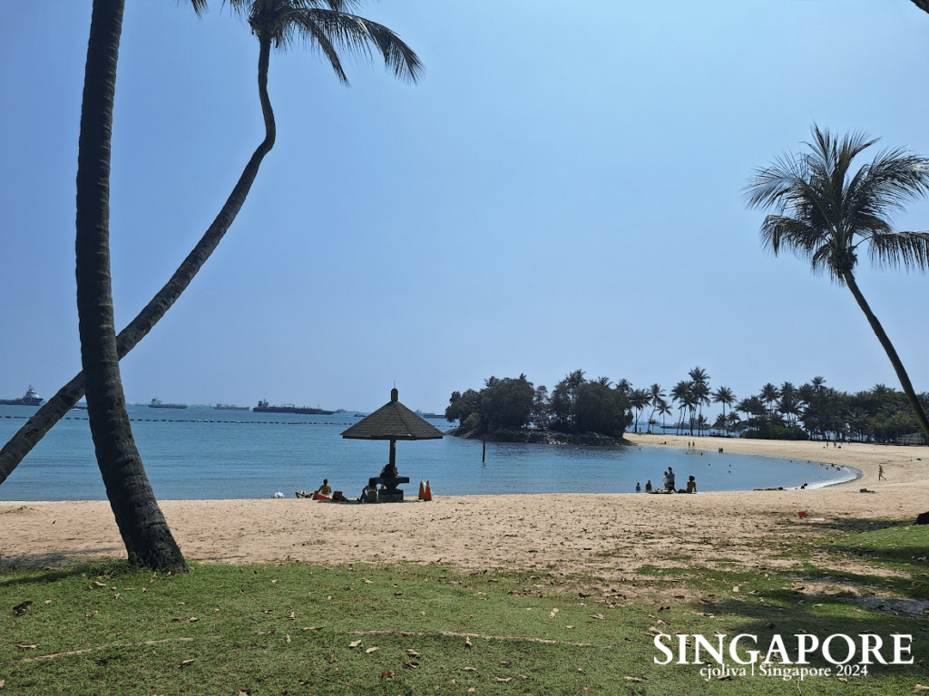 A scenic beach at Sentosa, Singapore, with golden sand, palm trees, a shaded hut, turquoise waters, and distant ships under a clear blue sky.