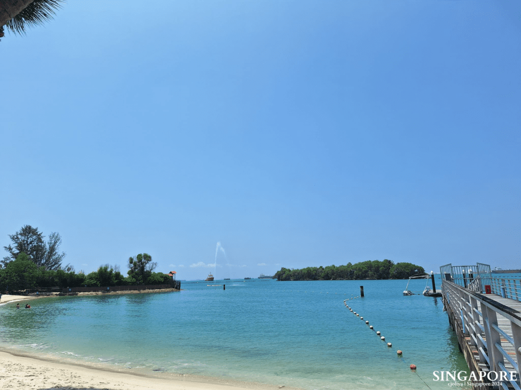 Scenic coastal view at Sentosa, Singapore, with a sandy beach, turquoise waters, a wooden pier, floating buoys, and a distant ship spraying water under clear blue skies.