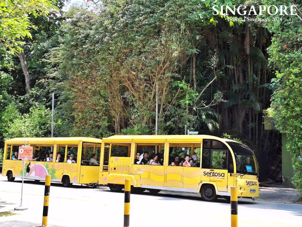 A bright yellow Sentosa tram with two carriages traveling along a paved road surrounded by lush tropical greenery in Singapore.