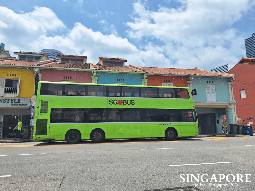 A bright green SG♥BUS double‑decker bus is parked along a Singapore street with colorful shophouses, shuttered windows, tiled roofs, and modern high‑rises in the background.