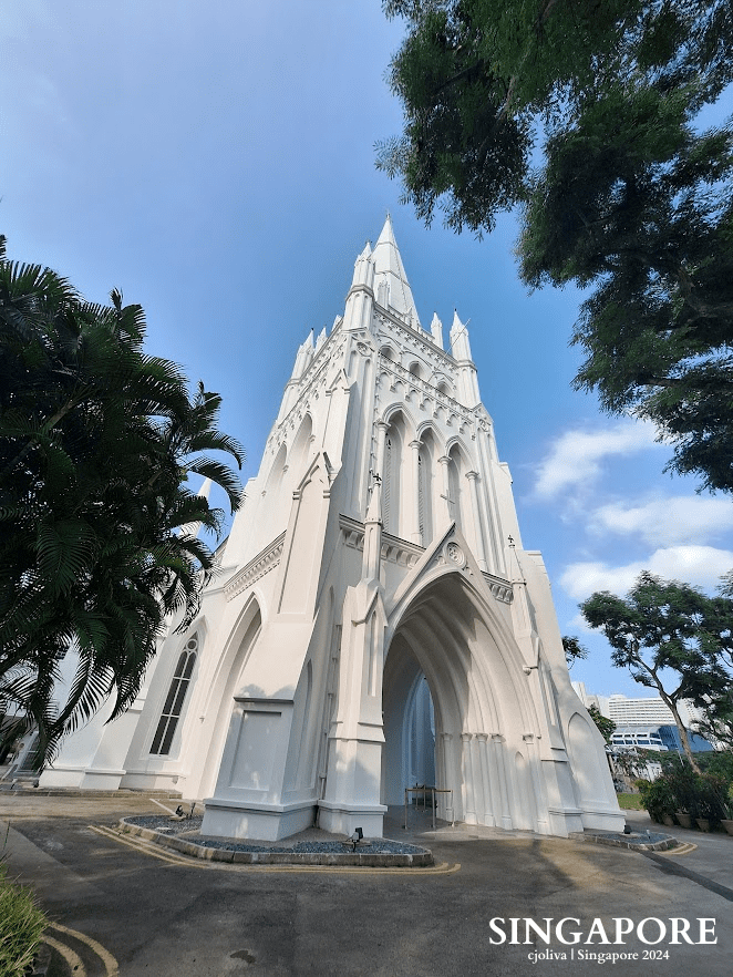 Tall white neo-Gothic St. Andrew’s Cathedral in Singapore with pointed arches, ribbed vaults, a central spire, and intricate detailing surrounded by greenery under clear blue skies.
