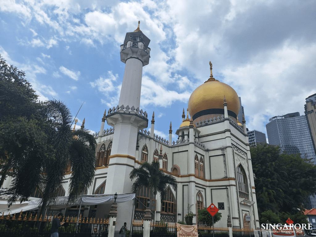 Sultan Mosque in Singapore with golden dome, white minaret, ornate arches, palm trees, and modern high-rises in the background.