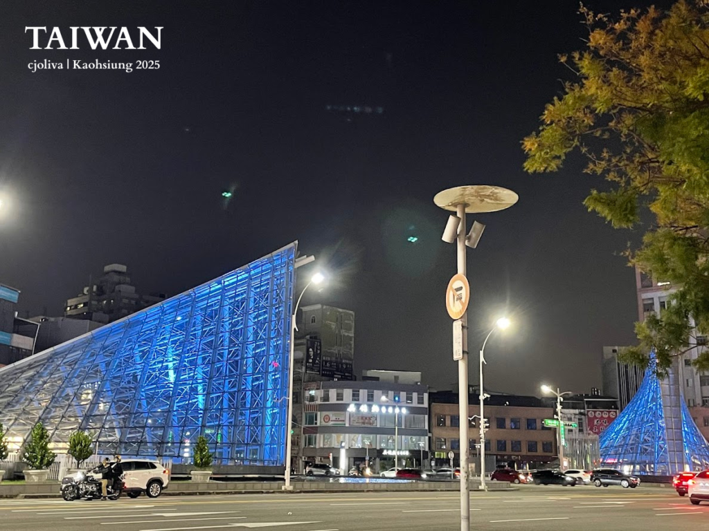 Exterior view of the illuminated blue glass pyramid structures of Formosa Boulevard MRT station in Kaohsiung at night, with city traffic and trees in the foreground.