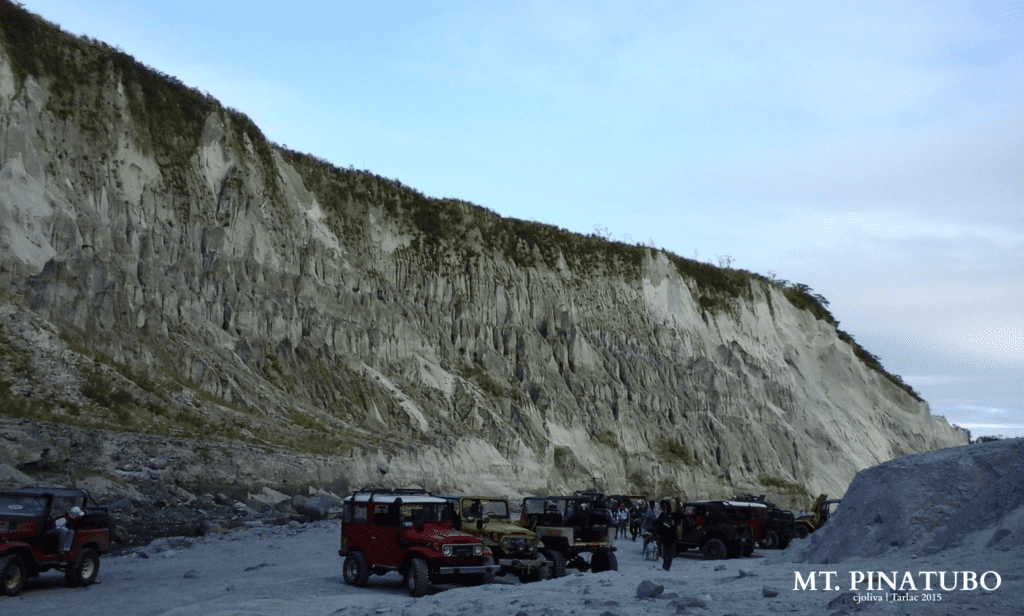 Several colorful jeepneys parked near towering volcanic cliffs at Mt. Pinatubo in Tarlac, with visitors walking around, 2015.
