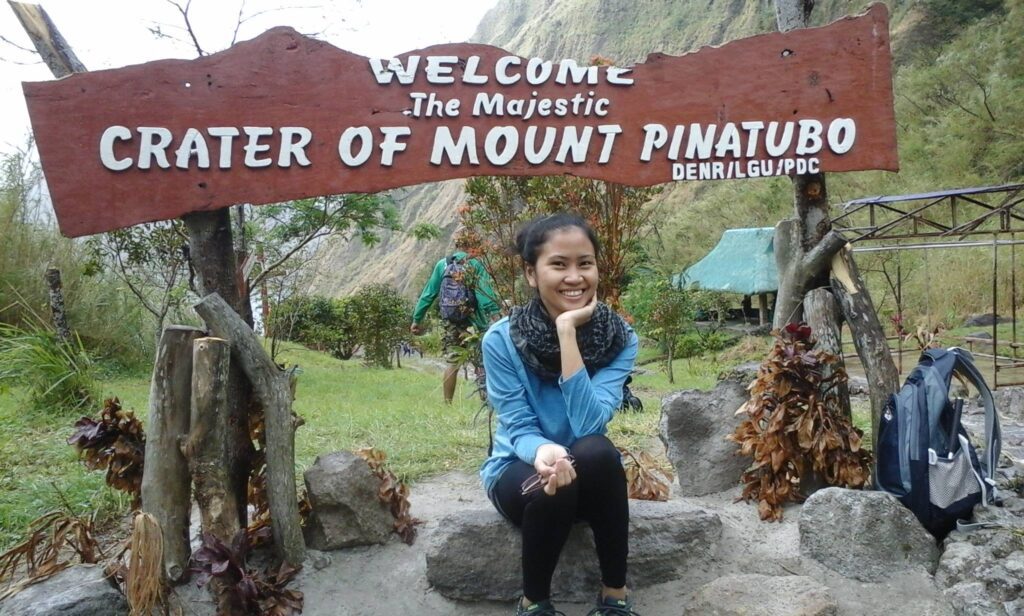 A traveler is sitting and smiling under a wooden welcome sign that reads "Welcome to The Majestic Crater of Mount Pinatubo."