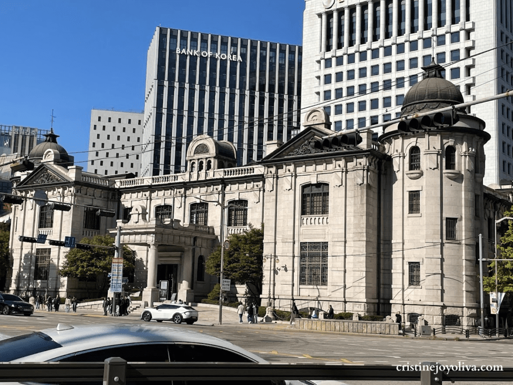 Daytime view of the historic Bank of Korea Money Museum in Seoul, South Korea. The Renaissance-style stone building with green domes is shown in the foreground, with the modern Bank of Korea skyscraper behind it.