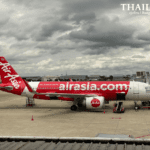A side profile of a red and white AirAsia Airbus A320 airplane parked at the gate under a cloudy sky at Don Mueang International Airport, Bangkok.