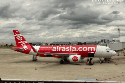 A side profile of a red and white AirAsia Airbus A320 airplane parked at the gate under a cloudy sky at Don Mueang International Airport, Bangkok.