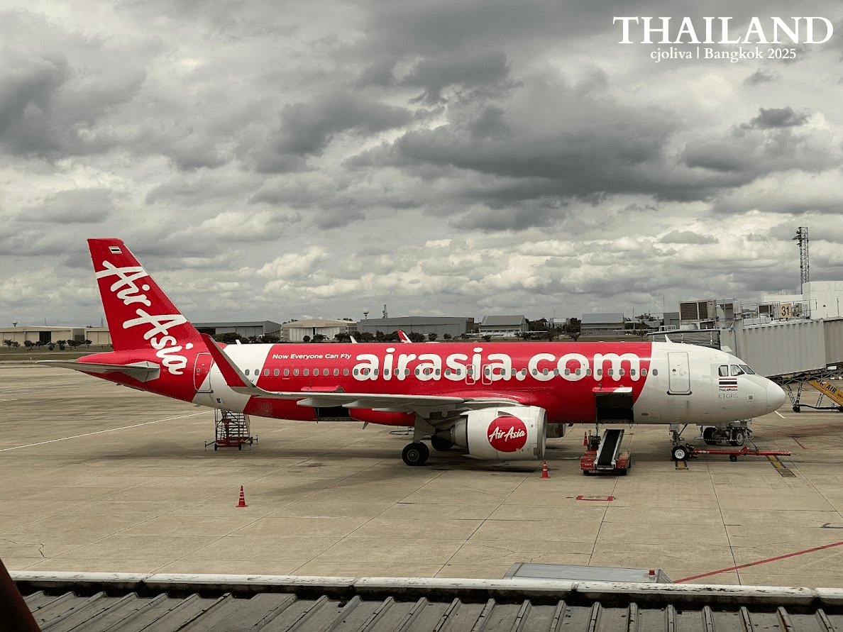 A side profile of a red and white AirAsia Airbus A320 airplane parked at the gate under a cloudy sky at Don Mueang International Airport, Bangkok.