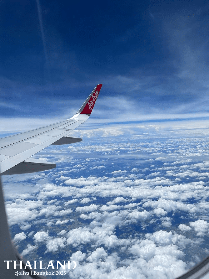 View from an airplane window showing an AirAsia wingtip over a dense field of white cumulus clouds under a deep blue sky during a flight to Thailand.