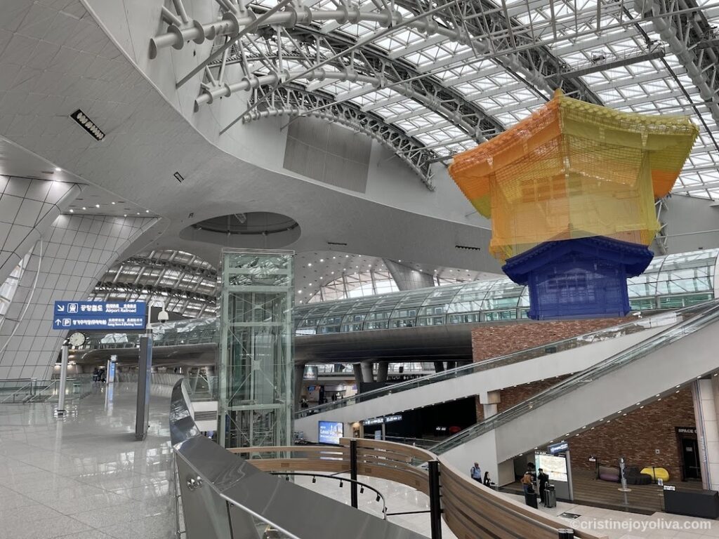 Interior of Korean transportation hub with curved glass ceiling, colorful suspended art installation resembling traditional building, escalators, and signage for Airport Railroad