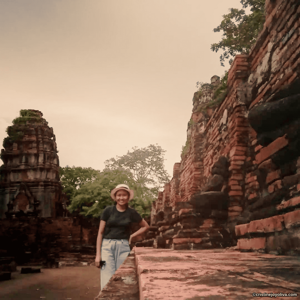 Person standing near ancient brick ruins in Ayutthaya, Thailand with weathered red walls, eroded statues, and a tall carved temple structure surrounded by greenery.