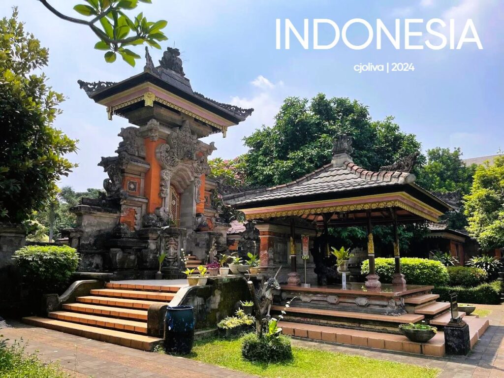 A sunny daytime view of a Balinese-style pavilion at TMII Jakarta, featuring an ornate orange-and-grey stone gateway with intricate carvings and a traditional open-air wooden gazebo (bale) with a tiered roof.
