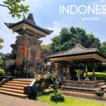 A sunny daytime view of a Balinese-style pavilion at TMII Jakarta, featuring an ornate orange-and-grey stone gateway with intricate carvings and a traditional open-air wooden gazebo (bale) with a tiered roof.
