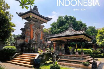 A sunny daytime view of a Balinese-style pavilion at TMII Jakarta, featuring an ornate orange-and-grey stone gateway with intricate carvings and a traditional open-air wooden gazebo (bale) with a tiered roof.