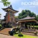 A sunny daytime view of a Balinese-style pavilion at TMII Jakarta, featuring an ornate orange-and-grey stone gateway with intricate carvings and a traditional open-air wooden gazebo (bale) with a tiered roof.