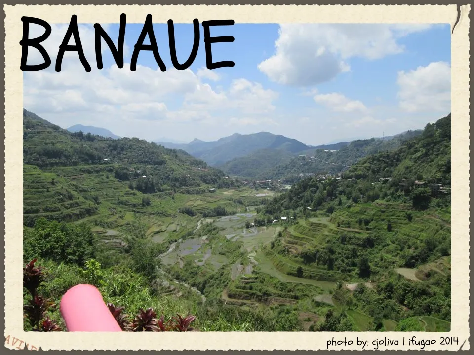 A panoramic view of the lush green Banaue Rice Terraces stretching across a wide valley with a river at the bottom, under a bright blue sky with scattered clouds.