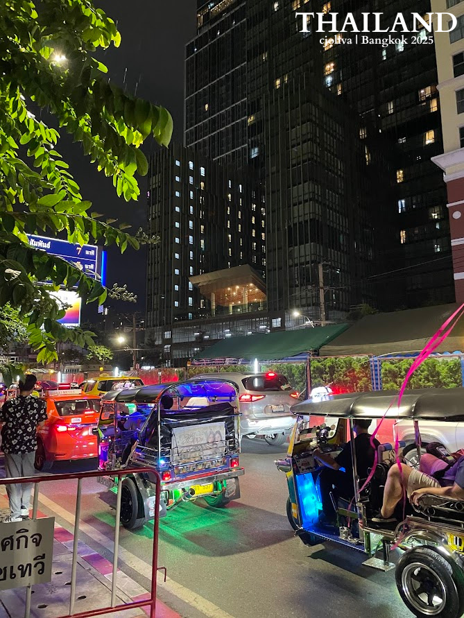 A nighttime street view in Bangkok, Thailand, featuring several tuk-tuks with neon lights stuck in traffic next to tall, modern buildings.