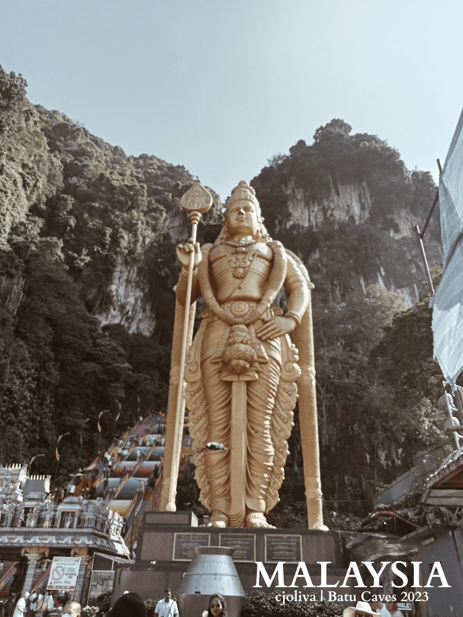 Golden Lord Murugan statue at Batu Caves in Malaysia, with colorful staircase leading to limestone caves and lush greenery in the background.