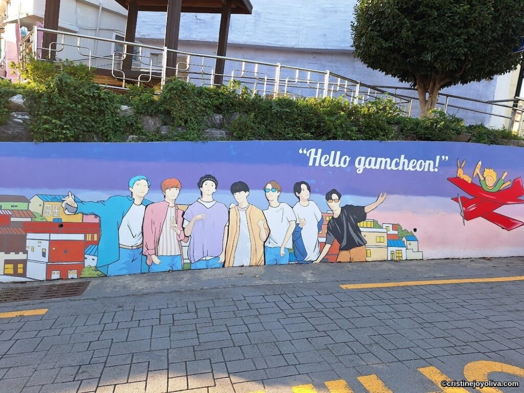 Wide shot of a large wall mural on a street in Gamcheon Culture Village, Busan, featuring stylized cartoon depictions of the seven members of BTS in various poses. To the right, the words 'Hello Gamcheon!' are in white text against a purple sky. The mural also shows stylized Gamcheon houses and a red airplane carrying characters. The mural is at street level on a paved sidewalk.