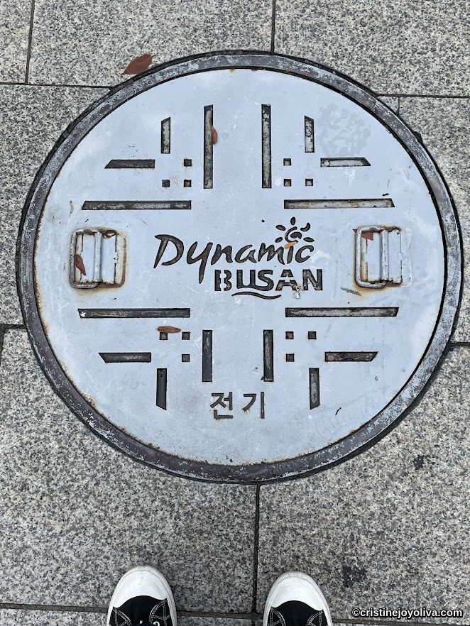 Dynamic Busan manhole cover with electricity symbol on tiled sidewalk
