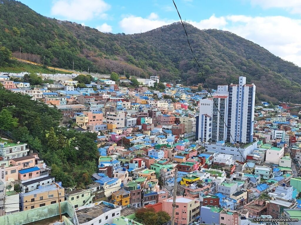 Wide-angle aerial view of Gamcheon Culture Village in Busan, South Korea, showing thousands of brightly painted houses tiered on a mountainside, with tall modern apartment buildings and a forested mountain in the background
