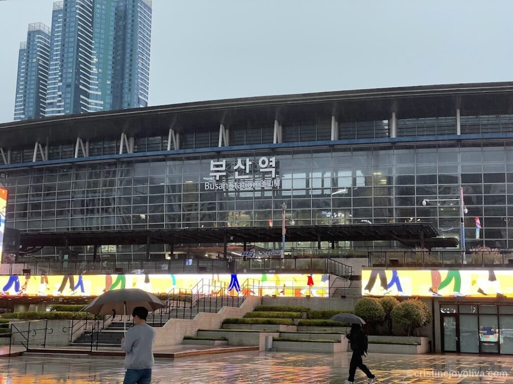 "Exterior of Busan Station with glass facade, multilingual signage, people walking with umbrellas on rainy day, and skyscrapers in background