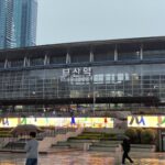 "Exterior of Busan Station with glass facade, multilingual signage, people walking with umbrellas on rainy day, and skyscrapers in background