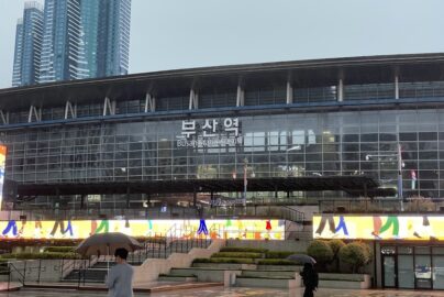 "Exterior of Busan Station with glass facade, multilingual signage, people walking with umbrellas on rainy day, and skyscrapers in background