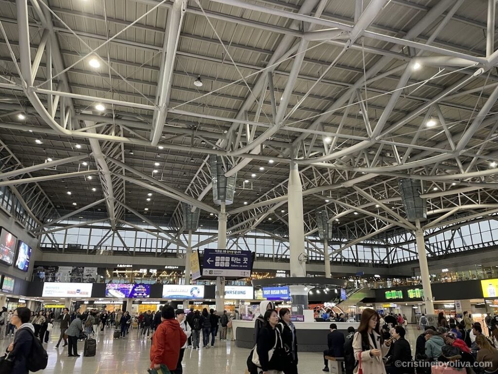 Interior of Busan Station with high steel ceiling, digital screens, directional signage, and travelers moving through spacious transit hub