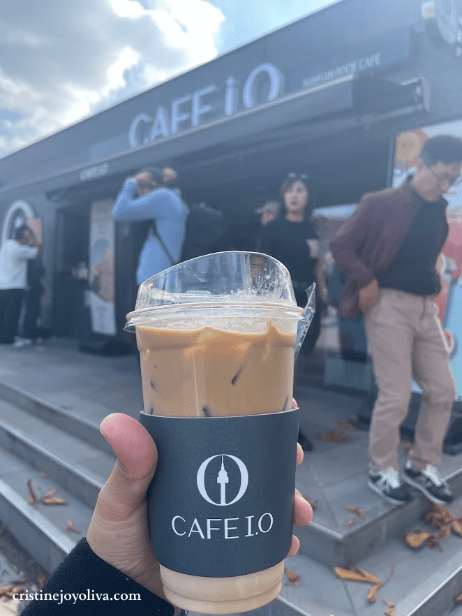 A hand holding an iced coffee from Cafe I.O with the Namsan Seoul Tower logo on the sleeve, standing in front of the cafe exterior at Namsan Park.