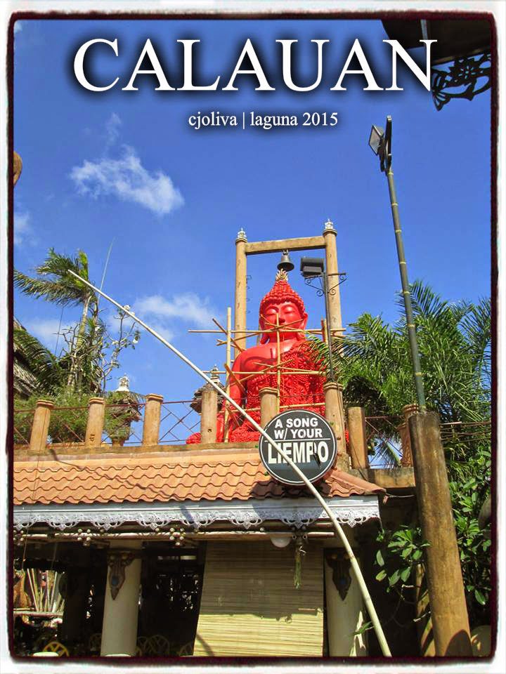Large red statue seated on a rooftop structure with decorative columns, palm trees, and tropical sky in Calauan, Laguna, Philippines
