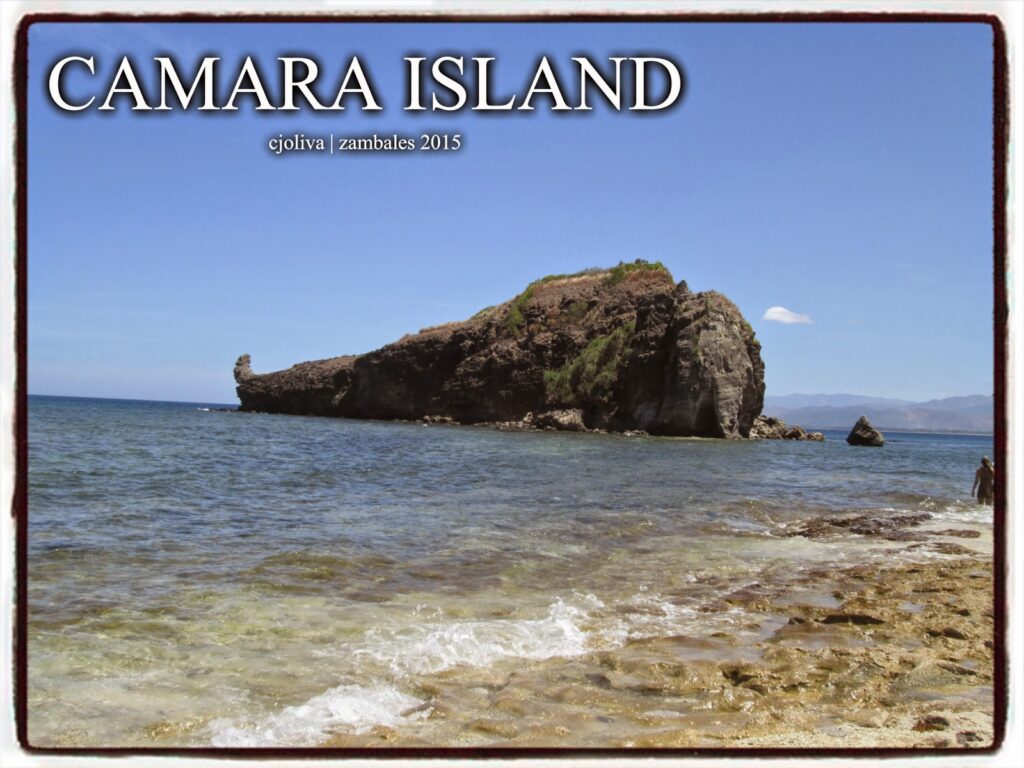 A rocky beach with clear, shallow waves in the foreground and a large, dark rock formation island under a clear blue sky in Zambales.